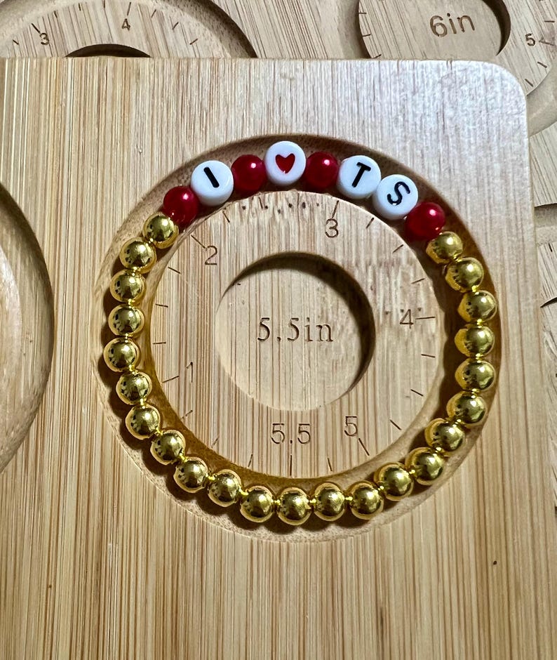 Op de afbeelding: Gouden kralenarmband met de letters "I LOVE TS" in witte en rode kralen. De armband wordt getoond op een houten meetinstrument met een cirkel met een diameter van 14 cm.
