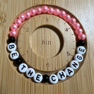 May include: A pink and black beaded bracelet with the words "BE THE CHANGE" spelled out on white beads. The bracelet is 6 inches in diameter and is laid out on a wooden surface.
