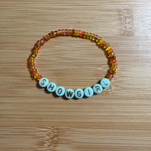 Puede incluir: Una pulsera de cuentas con la palabra "SHOWGIRL" escrita con cuentas de letras blancas. La pulsera tiene cuentas naranjas, amarillas y rosas, creando un accesorio colorido. La pulsera se muestra sobre una superficie de madera.