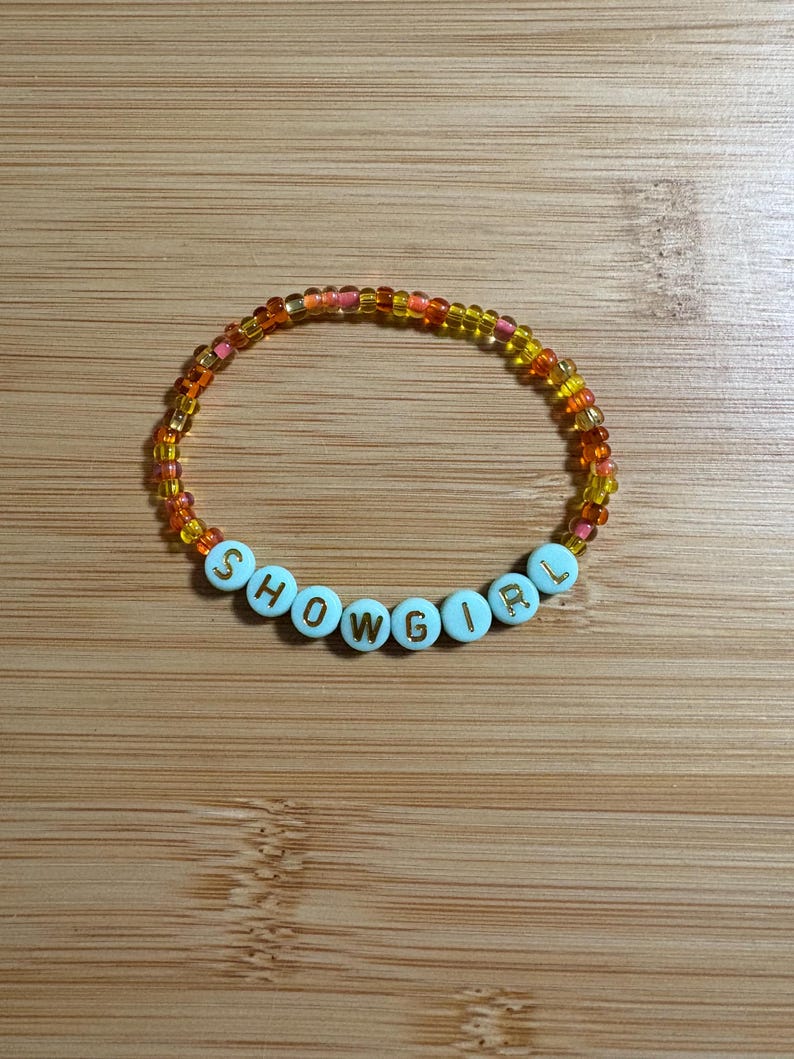 Puede incluir: Una pulsera de cuentas con la palabra "SHOWGIRL" escrita con cuentas de letras blancas. La pulsera tiene cuentas naranjas, amarillas y rosas, creando un accesorio colorido. La pulsera se muestra sobre una superficie de madera.