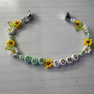 May include: A beaded bracelet with the words "OUR SONG" spelled out in white and gold letter beads. The bracelet features yellow and white flower beads, green leaf beads, and pearl beads. The bracelet has silver clasps.