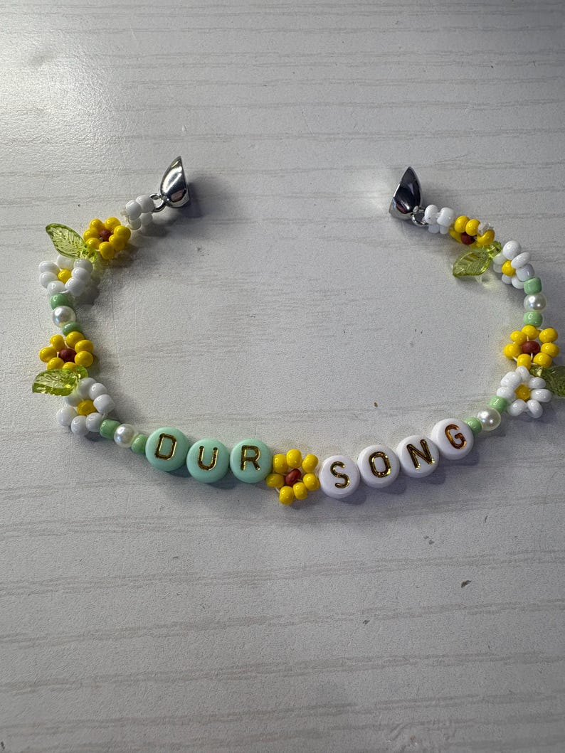 May include: A beaded bracelet with the words "OUR SONG" spelled out in white and gold letter beads. The bracelet features yellow and white flower beads, green leaf beads, and pearl beads. The bracelet has silver clasps.