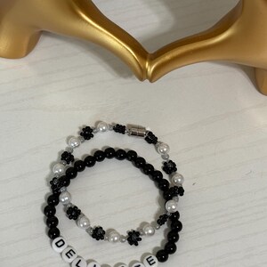 May include: Two beaded bracelets. One bracelet is black with white beads and black flower accents. The other bracelet is black with white beads and the word "DELICATE" spelled out in white letter beads. A gold heart-shaped sculpture is in the background.