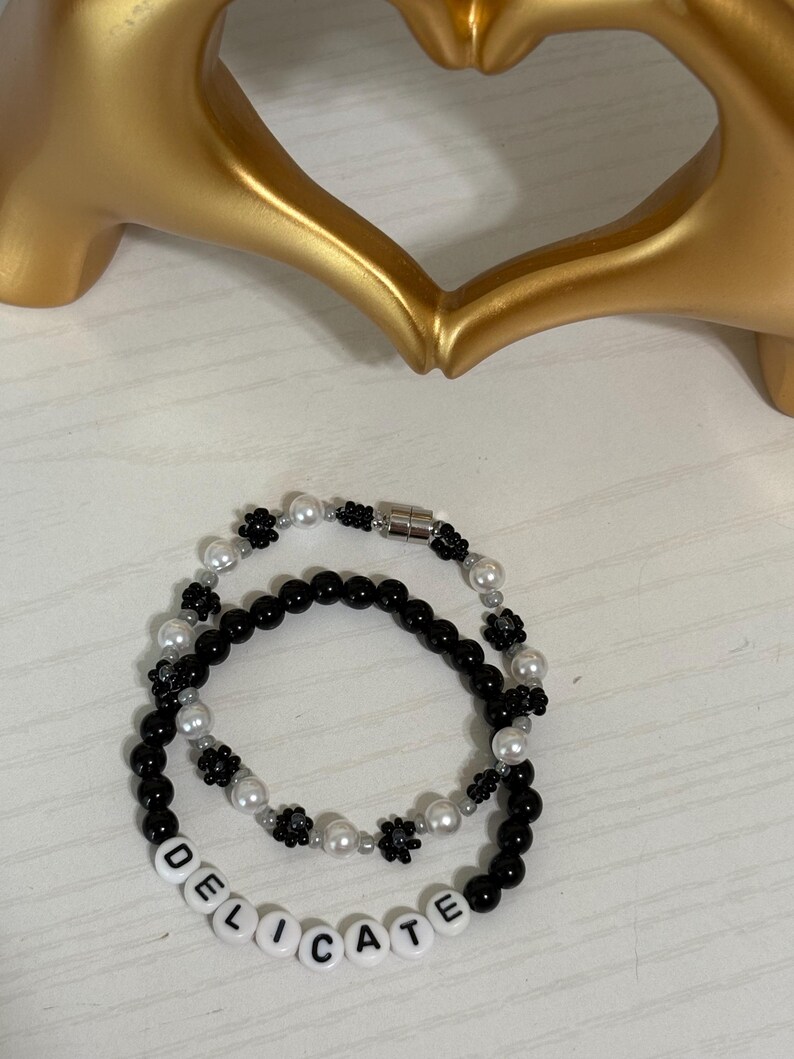 May include: Two beaded bracelets. One bracelet is black with white beads and black flower accents. The other bracelet is black with white beads and the word "DELICATE" spelled out in white letter beads. A gold heart-shaped sculpture is in the background.