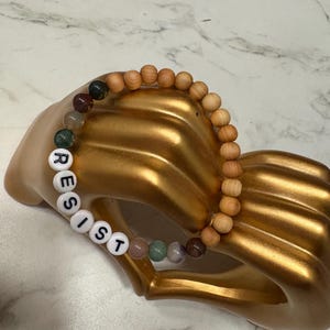 May include: A wooden beaded bracelet with the word "RESIST" spelled out in white beads. The bracelet also features a variety of colorful gemstone beads.