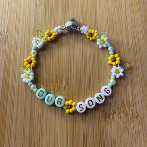 May include: A beaded bracelet with the words "OUR SONG" spelled out in white and light blue beads. The bracelet features yellow and white flower beads, green leaf beads, and a silver heart-shaped clasp. The bracelet is on a wooden surface.