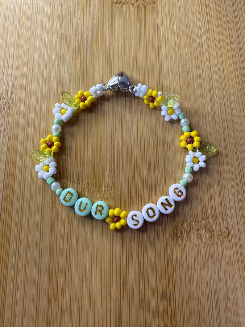 May include: A beaded bracelet with the words "OUR SONG" spelled out in white and light blue beads. The bracelet features yellow and white flower beads, green leaf beads, and a silver heart-shaped clasp. The bracelet is on a wooden surface.