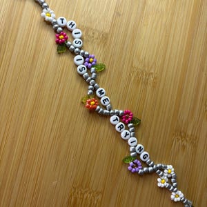 May include: A beaded bracelet spelling out "THIS TIME TRYING" with white letter beads. The bracelet has silver beads, colourful flower accents, and green leaf accents. The bracelet is displayed on a wooden surface.