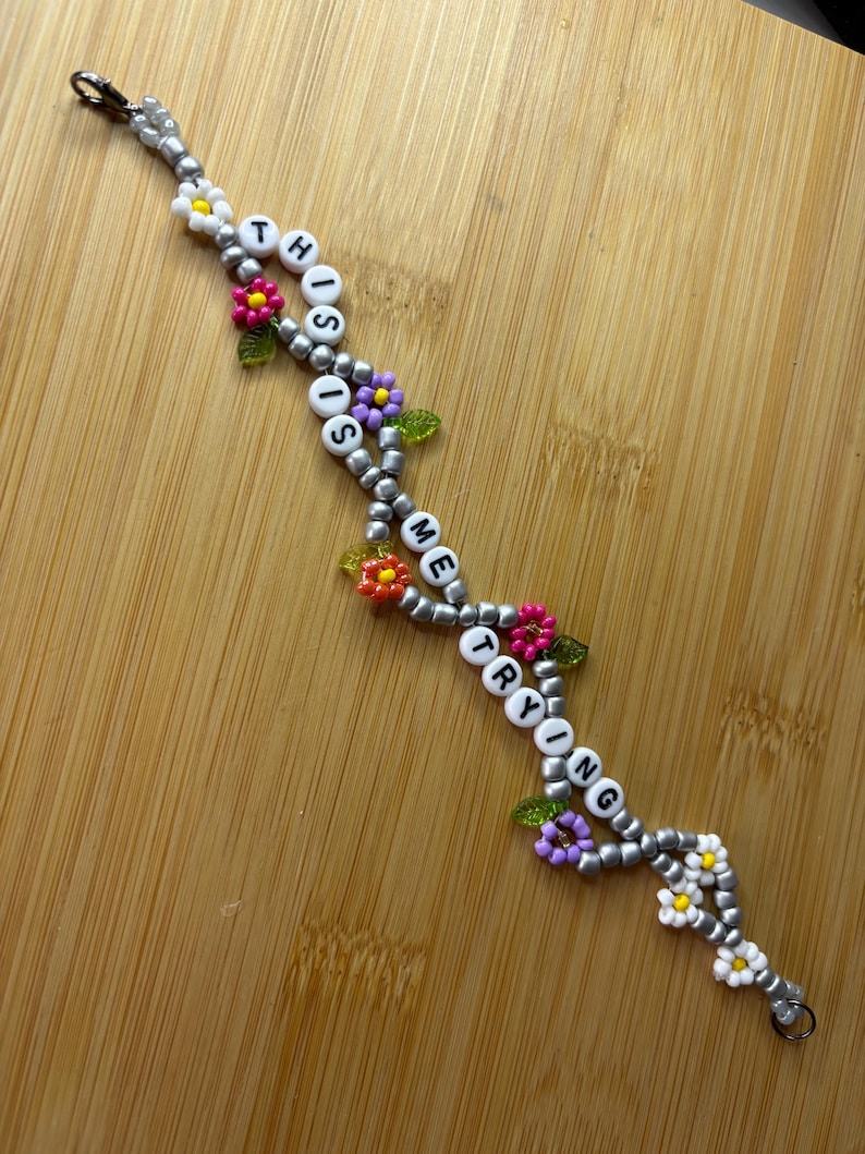 May include: A beaded bracelet spelling out "THIS TIME TRYING" with white letter beads. The bracelet has silver beads, colourful flower accents, and green leaf accents. The bracelet is displayed on a wooden surface.
