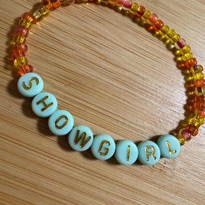 May include: A beaded bracelet with the word "SHOWGIRL" spelled out in gold letters on light blue beads. The rest of the bracelet is made of small, colorful beads in shades of yellow, orange, and pink.