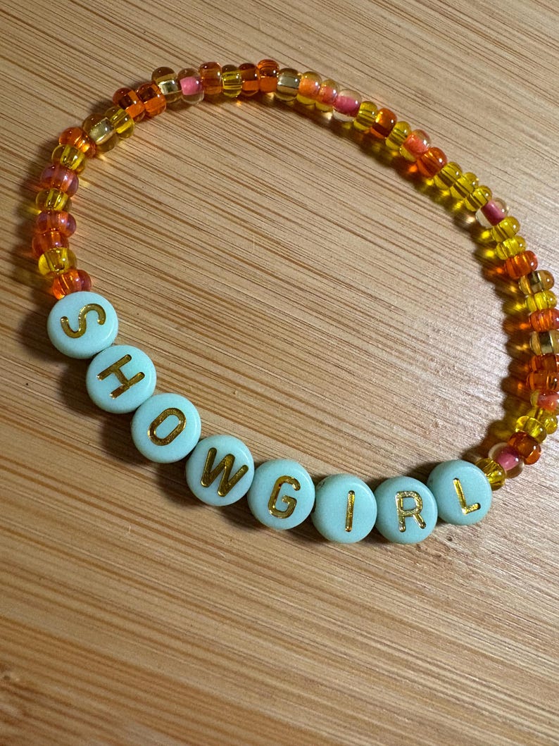 Puede incluir: Una pulsera de cuentas con la palabra "SHOWGIRL" escrita en letras doradas sobre cuentas azul claro. El resto de la pulsera est&aacute; hecho de peque&ntilde;as cuentas de colores en tonos amarillos, naranjas y rosas.
