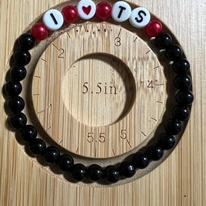 Op de afbeelding: Een zwart kralenarmband met witte letterkralen met de tekst "I ❤️ TS" en rode hart- en ronde kralen. De armband wordt gepresenteerd op een houten meetinstrument.