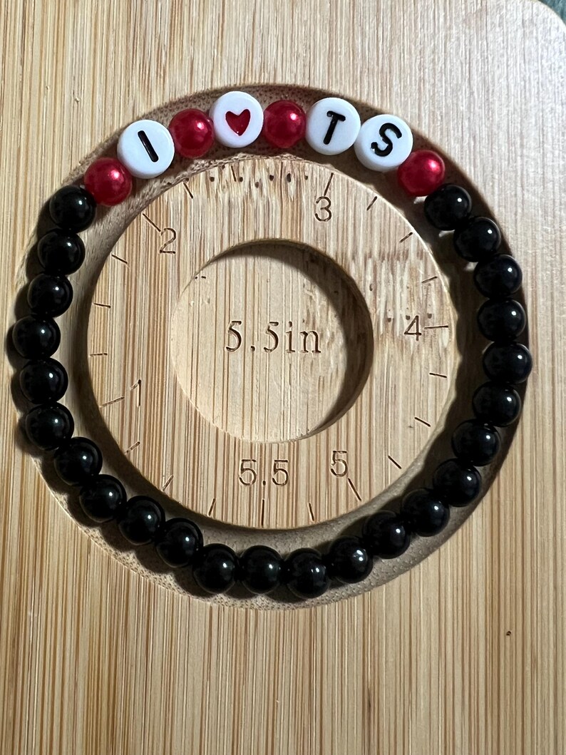 Op de afbeelding: Een zwart kralenarmband met witte letterkralen met de tekst "I ❤️ TS" en rode hart- en ronde kralen. De armband wordt gepresenteerd op een houten meetinstrument.