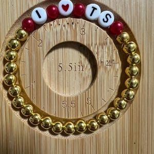 Op de afbeelding: Gouden kralenarmband met de letters "I ❤️ TS" in witte en rode kralen. De armband ligt op een houten meetinstrument met een diameter van ongeveer 14 cm.