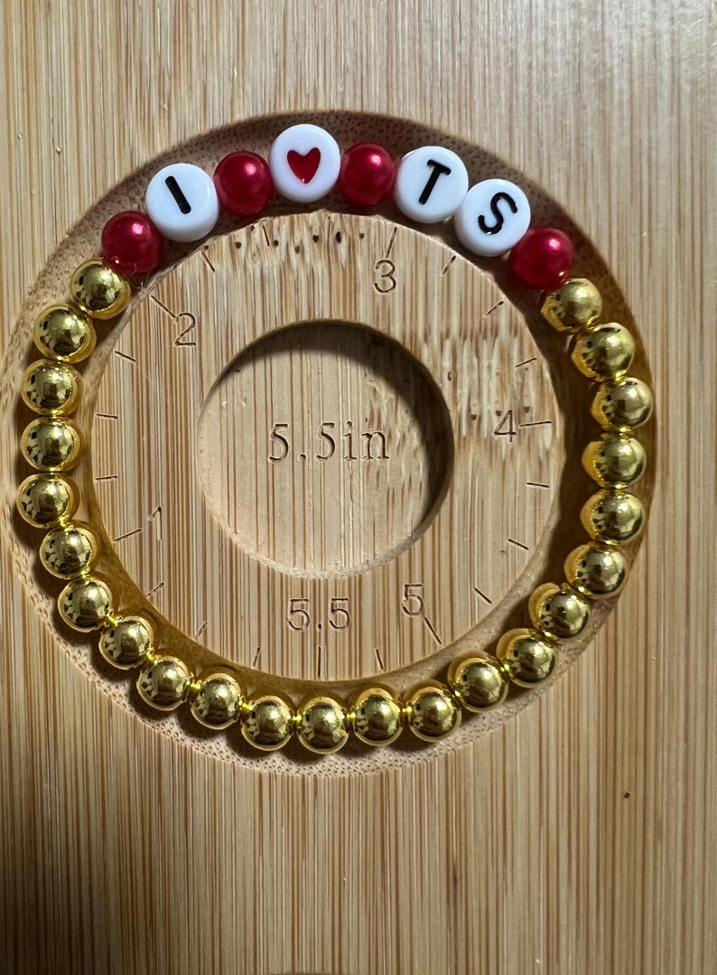 Op de afbeelding: Gouden kralenarmband met de letters "I ❤️ TS" in witte en rode kralen. De armband ligt op een houten meetinstrument met een diameter van ongeveer 14 cm.