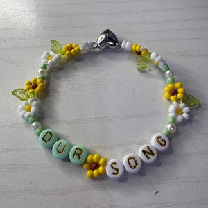 May include: A beaded bracelet with the words "OUR SONG" spelled out in white and mint green letter beads. The bracelet is decorated with yellow and white flower beads, green leaf beads, and white and yellow beads.