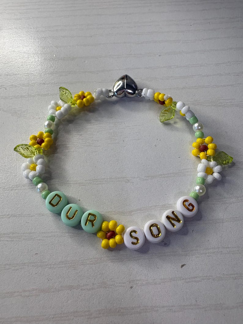 May include: A beaded bracelet with the words "OUR SONG" spelled out in white and mint green letter beads. The bracelet is decorated with yellow and white flower beads, green leaf beads, and white and yellow beads.