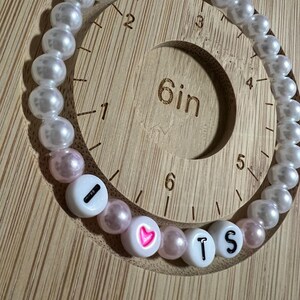 Op de afbeelding: Een kralenarmband met witte en roze kralen. De armband spelt "I ❤️ TS" met witte letterkralen en een roze hart. De armband wordt weergegeven op een houten meetinstrument.