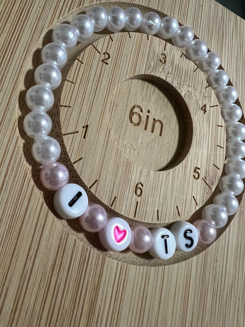 Op de afbeelding: Een kralenarmband met witte en roze kralen. De armband spelt "I ❤️ TS" met witte letterkralen en een roze hart. De armband wordt weergegeven op een houten meetinstrument.