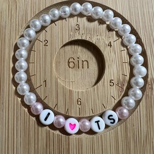 Op de afbeelding: Een kralenarmband met witte en roze parels. De armband spelt "I ❤️ TS" met witte letterkralen en een roze hartkralen. De armband is ongeveer 15 cm lang.