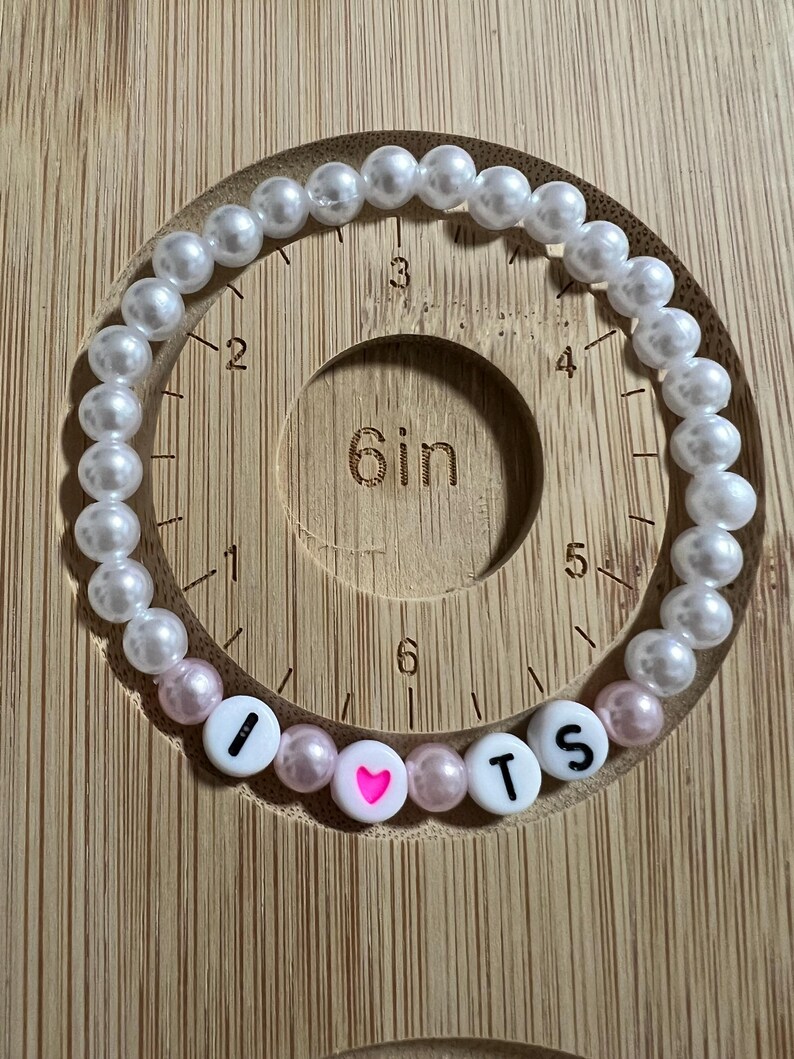 Op de afbeelding: Een kralenarmband met witte en roze parels. De armband spelt "I ❤️ TS" met witte letterkralen en een roze hartkralen. De armband is ongeveer 15 cm lang.