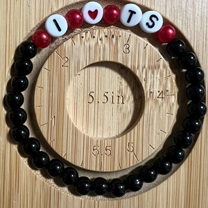 Op de afbeelding: Een kralenarmband met zwarte en rode kralen. De armband spelt "I ❤️ TS" met witte letterkralen en een rode hartkralen. De armband heeft een diameter van ongeveer 14 cm.