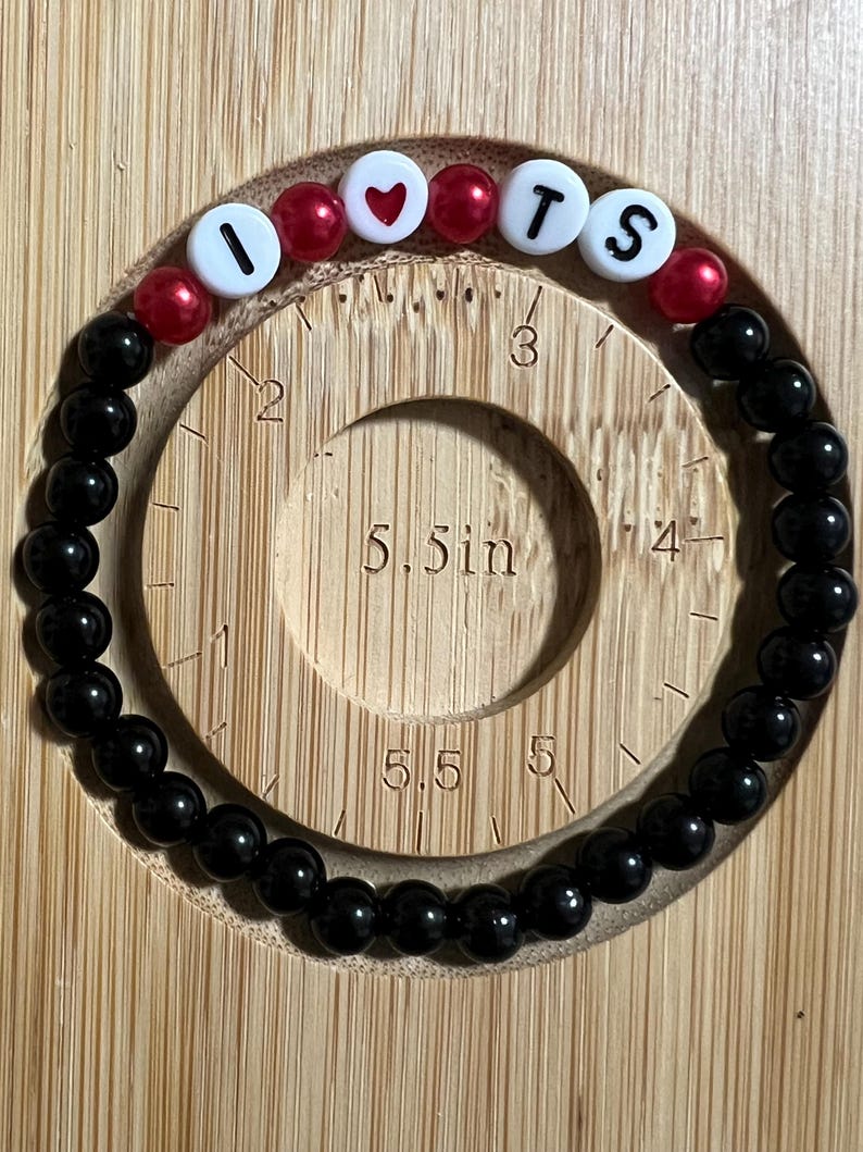 Op de afbeelding: Een kralenarmband met zwarte en rode kralen. De armband spelt "I ❤️ TS" met witte letterkralen en een rode hartkralen. De armband heeft een diameter van ongeveer 14 cm.