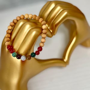 May include: A wooden bead bracelet with a mix of colorful gemstone beads. The bracelet is displayed on a gold-colored hand sculpture. The beads are various colors, including red, green, and white.
