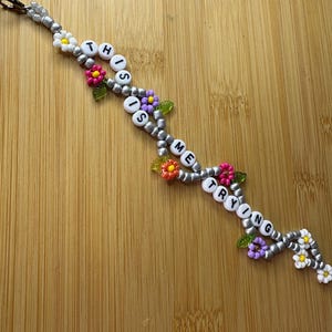 May include: A beaded keychain with the words "THIS IS ME TRYING" spelled out in white letter beads. The keychain is decorated with silver beads and colourful flower accents in pink, yellow, and purple.