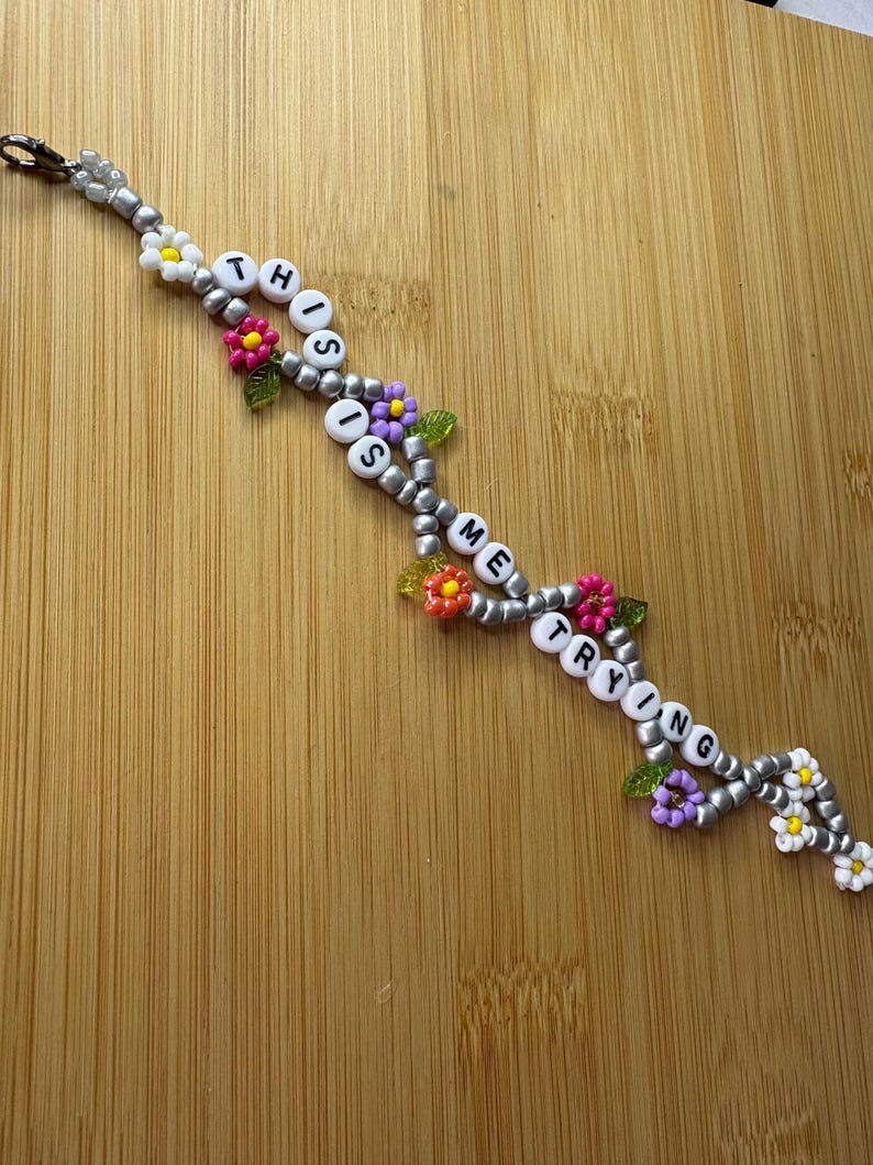May include: A beaded keychain with the words "THIS IS ME TRYING" spelled out in white letter beads. The keychain is decorated with silver beads and colourful flower accents in pink, yellow, and purple.