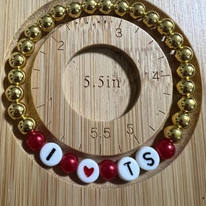 Op de afbeelding: Gouden kralenarmband met rode en witte accentkralen met de tekst "I ❤️ TS". De armband ligt op een houten meetinstrument. De gouden kralen hebben een diameter van ongeveer 0,6 cm.