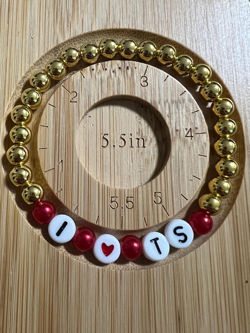 Op de afbeelding: Gouden kralenarmband met rode en witte accentkralen met de tekst "I ❤️ TS". De armband ligt op een houten meetinstrument. De gouden kralen hebben een diameter van ongeveer 0,6 cm.