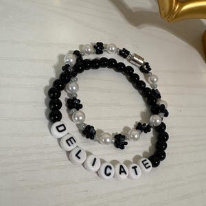 May include: Two beaded bracelets. One bracelet features black beads and white flower-shaped beads. The other bracelet has black and white beads, with the word "DELICATE" spelled out in white letter beads.