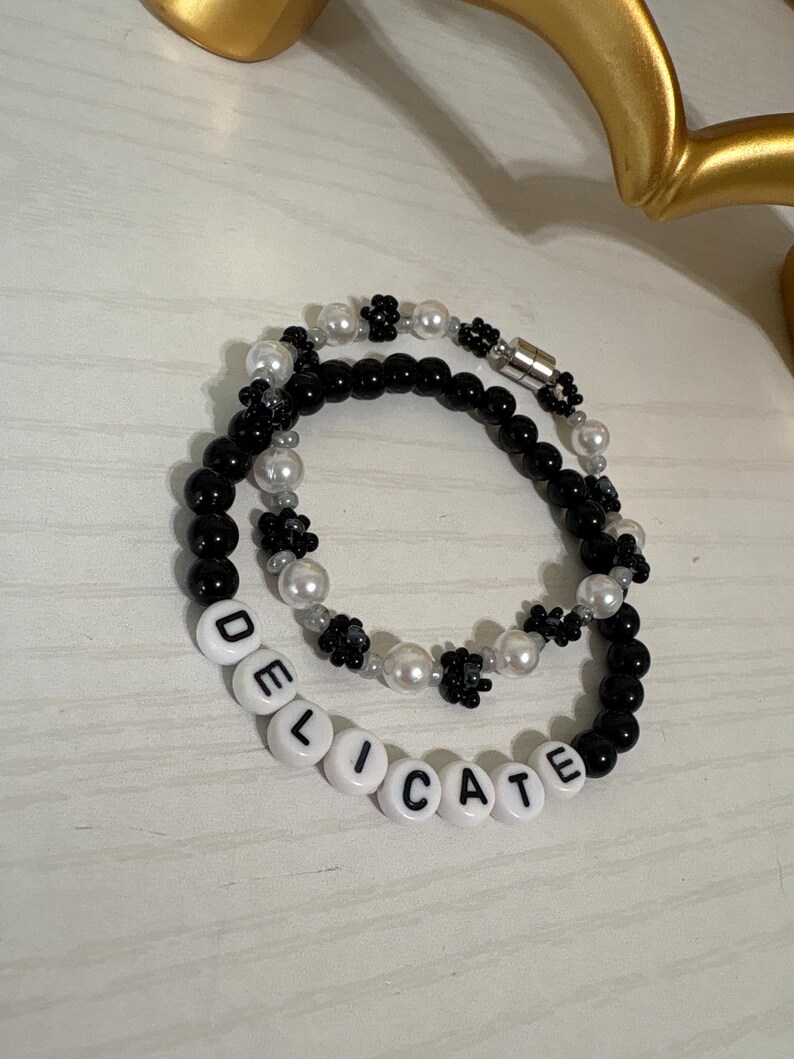 May include: Two beaded bracelets. One bracelet features black beads and white flower-shaped beads. The other bracelet has black and white beads, with the word "DELICATE" spelled out in white letter beads.