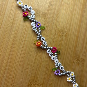May include: A beaded bracelet with the words "THIS IS ME TRYING" spelled out in white letter beads. The bracelet features silver beads, colourful flower accents, and a clasp.