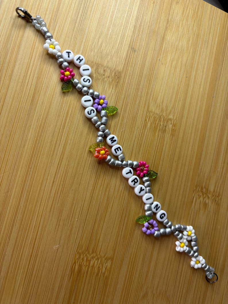 May include: A beaded bracelet with the words "THIS IS ME TRYING" spelled out in white letter beads. The bracelet features silver beads, colourful flower accents, and a clasp.