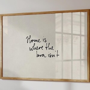 May include: A framed art print with the handwritten phrase "Home is where the bra isn't" in black script. The artwork is set against a neutral background and framed in light wood, creating a minimalist aesthetic.