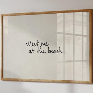 Puede incluir: Impresi&oacute;n enmarcada con el texto manuscrito "Meet me at the beach...". El marco es de madera clara y el fondo es de un beige neutro. La impresi&oacute;n se muestra en una pared blanca, con el reflejo de una ventana.