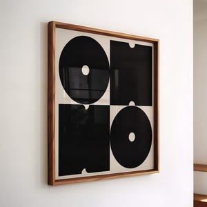 Può includere: Opera d'arte astratta in bianco e nero incorniciata. Il design presenta due cerchi e due quadrati neri su sfondo bianco. La cornice in legno offre un contrasto naturale e caldo. Appeso a una parete bianca.