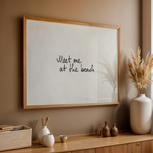 Puede incluir: Un cuadro enmarcado con el texto "Meet me at the beach" en escritura negra. El marco es de color marr&oacute;n claro, y el cuadro cuelga de una pared marr&oacute;n sobre un mueble de madera. Jarrones decorativos y una cesta est&aacute;n sobre el mueble.