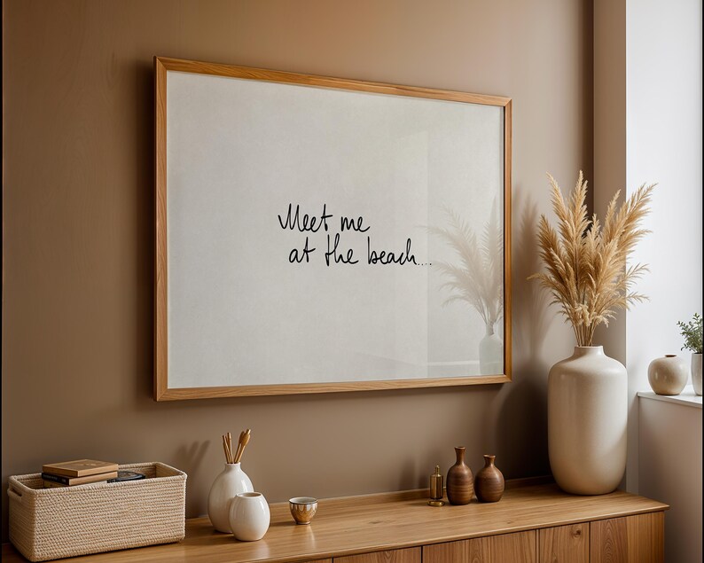 Puede incluir: Un cuadro enmarcado con el texto "Meet me at the beach" en escritura negra. El marco es de color marr&oacute;n claro, y el cuadro cuelga de una pared marr&oacute;n sobre un mueble de madera. Jarrones decorativos y una cesta est&aacute;n sobre el mueble.