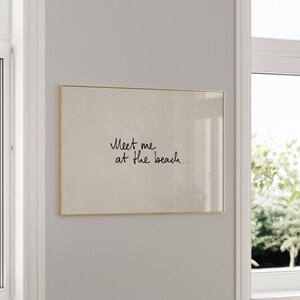 Puede incluir: Impresi&oacute;n rectangular enmarcada en dorado. La impresi&oacute;n presenta el texto manuscrito "Meet me at the beach..." en cursiva negra sobre un fondo beige. La obra enmarcada est&aacute; colgada en una pared gris claro.