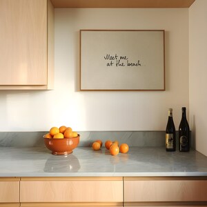 Puede incluir: Escena de cocina con un cuadro que dice "Meet me at the beach". Un cuenco de terracota lleno de naranjas descansa sobre una encimera gris, con m&aacute;s naranjas esparcidas. Dos botellas de vino tambi&eacute;n est&aacute;n presentes.