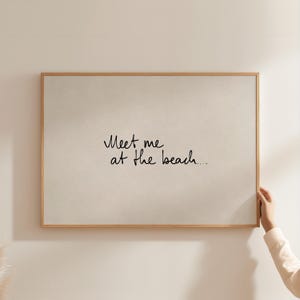 Puede incluir: Una impresi&oacute;n enmarcada con el texto "Meet me at the beach..." en escritura negra. La impresi&oacute;n tiene un fondo beige claro y un marco de madera natural. La imagen est&aacute; sostenida por una mano.