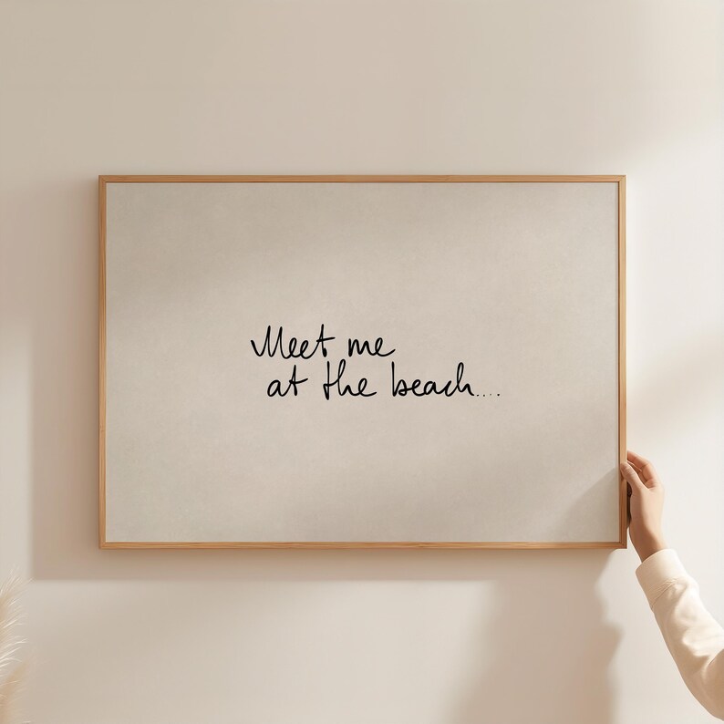 Puede incluir: Una impresi&oacute;n enmarcada con el texto "Meet me at the beach..." en escritura negra. La impresi&oacute;n tiene un fondo beige claro y un marco de madera natural. La imagen est&aacute; sostenida por una mano.