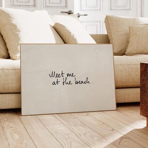 Puede incluir: Una impresi&oacute;n art&iacute;stica rectangular enmarcada con el texto manuscrito "Meet me at the beach..." en escritura negra. La impresi&oacute;n tiene un fondo blanco roto neutro y un marco dorado fino. La obra de arte est&aacute; apoyada contra un sof&aacute; beige sobre un suelo de madera clara.