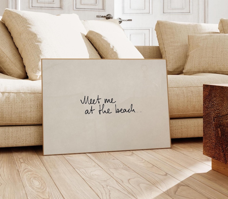 Puede incluir: Una impresi&oacute;n art&iacute;stica rectangular enmarcada con el texto manuscrito "Meet me at the beach..." en escritura negra. La impresi&oacute;n tiene un fondo blanco roto neutro y un marco dorado fino. La obra de arte est&aacute; apoyada contra un sof&aacute; beige sobre un suelo de madera clara.