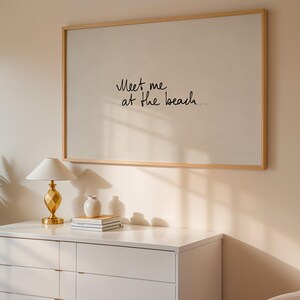 Puede incluir: Una obra de arte enmarcada con el texto "Meet me at the beach..." en escritura negra. La obra est&aacute; montada sobre una pared de color claro, encima de una c&oacute;moda blanca. Una l&aacute;mpara dorada, un peque&ntilde;o jarr&oacute;n blanco y libros est&aacute;n sobre la c&oacute;moda.