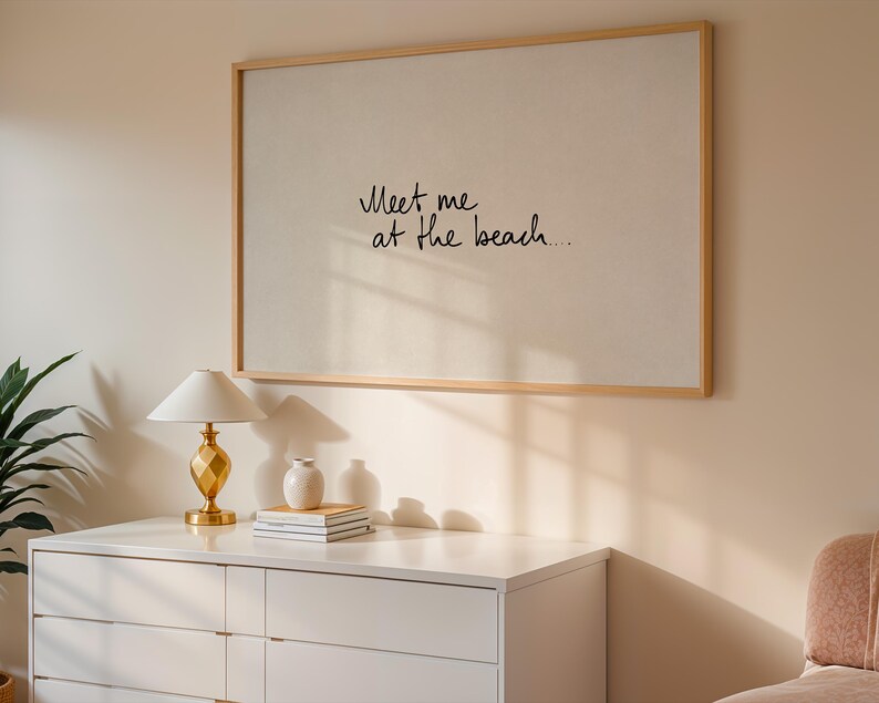 Puede incluir: Una obra de arte enmarcada con el texto "Meet me at the beach..." en escritura negra. La obra est&aacute; montada sobre una pared de color claro, encima de una c&oacute;moda blanca. Una l&aacute;mpara dorada, un peque&ntilde;o jarr&oacute;n blanco y libros est&aacute;n sobre la c&oacute;moda.