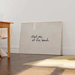 Puede incluir: Un cuadro enmarcado con el texto "Meet me at the beach..." en estilo manuscrito. El marco es dorado y el fondo es de color beige claro. El cuadro est&aacute; apoyado contra una pared blanca.